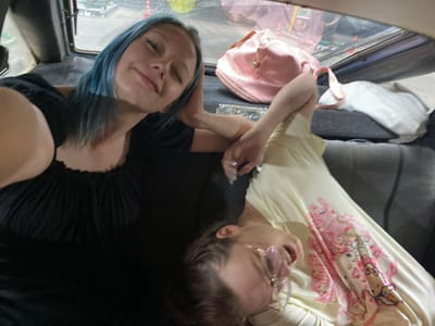 This is a selfie taken inside a vehicle, likely a car, showing two people. The person in the foreground on the right has blue hair and is looking directly at the camera with a slight smile. They are wearing a black top. The second person is on the left, wearing glasses and a light-colored t-shirt with a pink Hello Kitty graphic. Their mouth is open, as if speaking or laughing, and they are looking to the side. A pink duffel bag is visible between them. The setting appears to be casual, possibly a road trip or outing, with a dark interior of the vehicle and reflections visible on the windshield. The lighting suggests it could be daytime. The overall mood seems relaxed and friendly.