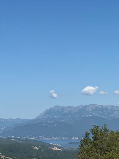 An eye-level, wide shot captures a vista of mountains, water, and foliage under a clear blue sky in Bratešići, Montenegro. In the foreground, lush green trees and bushes dominate the lower right quadrant, their leaves detailed and vibrant. Further back, rolling hills covered in dense green forests lead the eye towards a body of water that appears to be a bay or a large river. Along the distant shoreline, a town with white buildings is visible, nestled against the foothills. Dominating the background are several layers of rugged, blue-grey mountains, their rocky textures and ridges clearly defined. A few small, fluffy white clouds dot the otherwise uninterrupted expanse of the sky. The light suggests it is daytime, likely mid-morning or early afternoon, given the bright illumination and the clear visibility of the landscape. There are no people or animals visible in the frame.