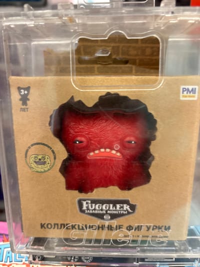 A red, fuzzy toy monster with large, droopy eyes and a wide, toothy grin is displayed in a clear plastic package. The packaging features a cardboard backing with a brick pattern and a logo that reads "Fugglers" above text in Russian that translates to "Funny Monsters." Below the monster's image, more Russian text on the cardboard reads "Collectible Figures." To the left of the monster, a circular emblem contains a depiction of a small, yellow creature. A "3+" age rating and a silhouette of a teddy bear are also visible on the packaging. The toy is located on a retail shelf, as suggested by the presence of other merchandise and the overall store-like environment. The specific location within the city of Kuzmolovsky, Russia, cannot be determined from the image alone.