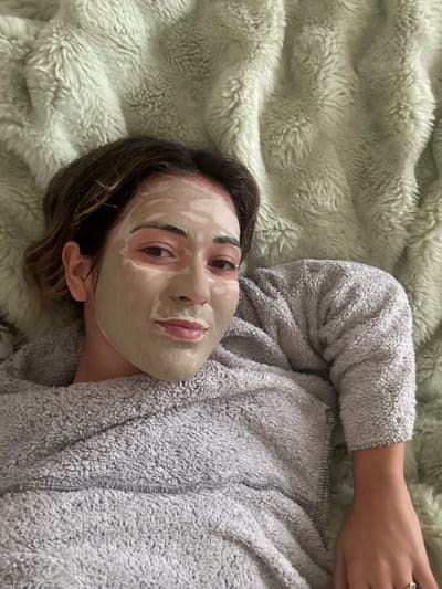 A female individual is depicted in a close-up, top-down perspective, lying supine indoors in the city of Kalyvia Thorikou Municipal Unit, Greece. Her face is covered with a light green facial mask, leaving the eyes, eyelids, under-eye areas, and lips exposed. Dark brown hair frames her forehead. She is wearing a grey, textured, long-sleeved fuzzy garment. A silver-colored ring with a clear stone is visible on the ring finger of her right hand. The individual makes direct eye contact with the camera, displaying a slight smile. She is positioned against a light green, shaggy-textured blanket that forms the entire background.