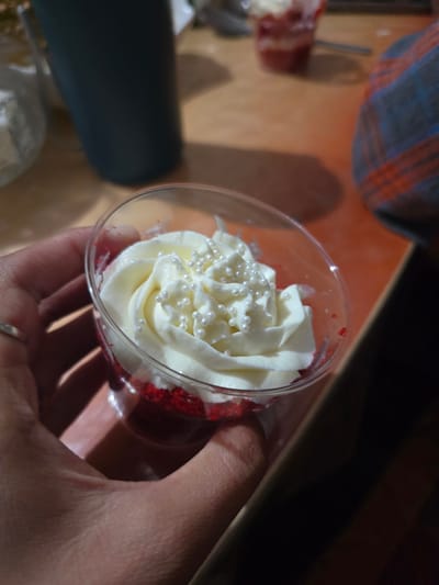 A close-up shot shows a hand holding a clear plastic cup containing a red velvet cupcake topped with white frosting and pearl sprinkles. The background is blurred, featuring a dark blue tumbler on the left and a plaid fabric on the right. The surface appears to be a wooden table. The scene is indoors, likely a domestic setting, and there is no indication of Boz Beshik, Kyrgyzstan.