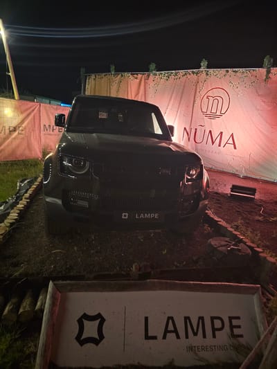 The image shows a black Land Rover Defender parked outdoors at night, possibly at an event. Behind the vehicle is a large, light-colored fabric banner with red lettering. The lettering on the banner includes the word "NUMA" and the phrase "SUMMERBAR". To the left of the Defender, another banner is partially visible with the text "MPE" and "INTERESTING CARS". The ground appears to be dirt and grass, with wooden logs used as a border in the foreground. The overall lighting suggests it is nighttime, with artificial lights illuminating the scene. The vehicle has its headlights on. The image was taken in Oudenaarde, Belgium.