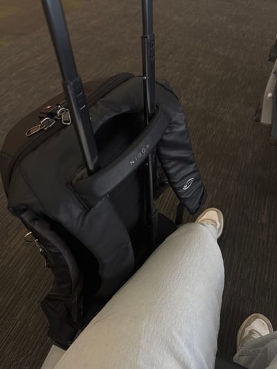 A first-person perspective shot shows a person's legs and a black backpack in what appears to be an airport terminal or a similar waiting area. The person is wearing light-colored denim jeans and white sneakers with light-brown accents. The backpack, which has a telescopic handle extended, is positioned to the left of the frame. The handle has a black grip with the brand name "KORIN" printed in white letters. A small circular logo is also visible on the grip. The background consists of a dark-colored carpet with a subtle striped pattern, characteristic of airport flooring. The lighting is ambient, suggesting an indoor setting. The overall scene captures a moment of waiting, possibly before a flight. The location is identified as Apodaca, Mexico.