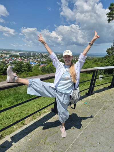 A smiling woman in a white baseball cap and a white unbuttoned shirt over a light blue t-shirt and loose light blue pants strikes a pose with her arms raised in a peace sign and one leg extended backwards, resting on a wooden railing. She is wearing pink athletic shoes and a patterned crossbody bag. The background shows a sprawling city with a mix of residential buildings and some industrial structures, nestled in a valley with green hills and mountains in the distance under a bright blue sky with scattered clouds. The scene is an outdoor viewpoint overlooking the city of Arseniev, Russia. The foreground consists of a paved walkway with patches of grass and a metal railing.