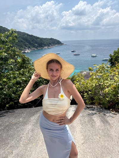 A woman stands on a paved surface near a lush green hillside and an ocean view in Lam Kaen, Thailand. She is wearing a straw hat, a white top with a flower, and a light blue skirt. She poses with one hand holding the brim of her hat and the other on her hip. The ocean is visible in the background, dotted with several boats.