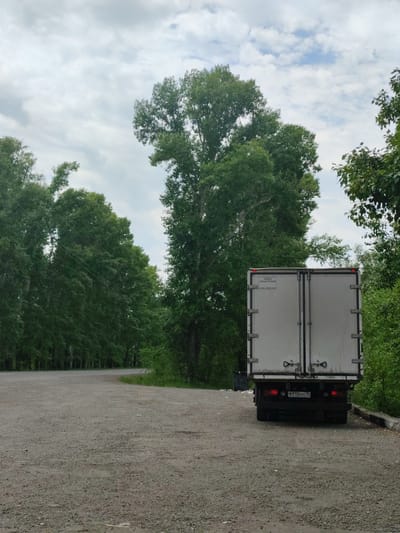 This media file depicts the rear of a white box truck parked on a gravel surface. The truck has a license plate that reads "B 715KH 19". Above the license plate, there is text on the truck door that says "ЗАВОД АВТОФУРГОНОВ" and "8-800-2000-424".
The truck is positioned in an outdoor setting with a backdrop of dense green trees on both sides of a curved road. The sky is overcast with clouds. The ground is covered with gravel and small stones. To the left of the truck, a black trash bin is visible. The scene appears to be a quiet, possibly rural area, given the natural surroundings and the type of road. There are no people visible in the media file. The time of day seems to be daytime, given the visibility, and the weather appears to be cloudy. The location cues of trees and road suggest a natural or semi-developed area.