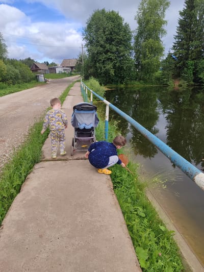 Two young boys are on a concrete path next to a body of water in Советско-Никольское, Russia. The boy on the left, facing away from the camera, is wearing a grey pajama set with a cartoon pattern and yellow shoes. He is standing near a stroller. The boy on the right is crouched down, facing the water, and holding a small orange net, seemingly trying to catch something in the shallow water near the grassy edge. The environment consists of a dirt road to the left, with houses and trees visible in the background, and a calm, reflective body of water to the right, bordered by a blue and white railing. The sky is partly cloudy.