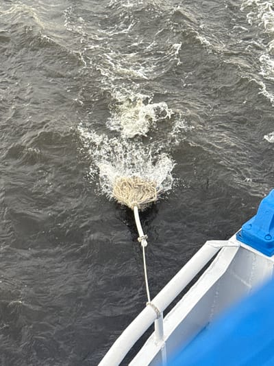 A mop is being dragged through dark, choppy water, creating a spray and wake. A white rope connects the mop to a white railing with a blue fixture attached. The scene is likely on the Neva River in the city of Belous, Russia, possibly from the deck of a boat.