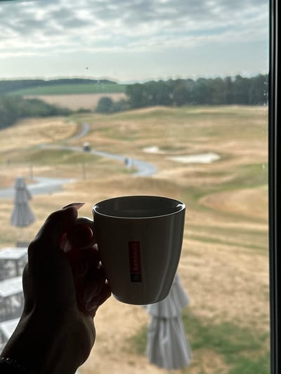 A person's hand, with manicured nails, holds a white ceramic mug with a red logo that says "Rombouts" against a blurred background of a golf course. The background shows green fairways, sand traps, a winding path, and a line of trees in the distance under a cloudy sky. The image is shot through a window, with reflections visible on the glass. The location is Méan, Belgium.