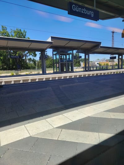 The media file depicts a view of a train station platform in Günzburg, Germany. The scene is set outdoors under a clear blue sky, suggesting it is daytime with good weather. The platform is paved with grey and light-colored rectangular tiles, with a diagonal shadow cast across it. In the background, there is a covered waiting area with a modern design, featuring glass walls and a metal roof. Several signs are visible, including a prominent blue sign that reads "Günzburg" in white lettering, indicating the station's name. Other signs with numbers and letters, such as "5", "D 4", and "405.0-F", are also present, likely referring to platforms or track information. Behind the covered area, there are trees and some buildings, including a glimpse of a white truck. The overall impression is of a quiet, functional public space on a bright day. There are no people visible in the image, emphasizing the emptiness of the platform. The image appears to have been taken from inside a train or from a similar vantage point, looking out towards the platform.