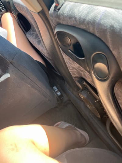 A close-up shot from inside a vehicle shows the legs of a person sitting in the passenger seat, next to the car door. The car's interior includes a black fabric seat, a black plastic door panel with a speaker and handle, and a beige trim. The person is wearing a dark skirt and light-colored sandals. The scene is illuminated by bright sunlight, casting shadows. The image's perspective is low, focusing on the lower body and interior elements. The location is identified as Слабодкаўскі сельскі Савет, Belarus.