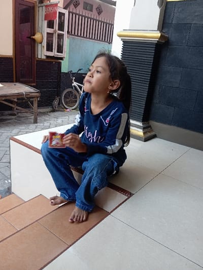 In the city of Pasuruan, Indonesia, a young female, estimated to be 6-10 years old, is seated barefoot on a light-colored tiled step. She has dark hair styled in a ponytail, wears small gold-colored earrings, a dark blue long-sleeved shirt with white arm stripes, and faded blue denim jeans. Her toenails are painted pink. She holds a small, rectangular red-orange packet in her left hand and gazes upwards and slightly to her left with a neutral expression.
The immediate ground consists of light beige tiles with dark brown trim for the step she occupies, and terracotta-colored tiles on the lower step where her feet rest. To the left, a paved outdoor area is visible, featuring a light-colored wooden table with bamboo-style legs. A dark brown wooden door is partially open, next to which a small wooden cabinet is mounted on the wall. A red sign with white text, reading "BENSIN" (gasoline), hangs from the cabinet, accompanied by a yellow funnel, indicating a roadside fuel vendor. A white bicycle is parked further back, and a small, dark-colored cat is observed near its front wheel. To the right of the subject, a decorative pillar with black horizontal tiles and gold-colored trim stands beside a wall composed of dark gray/black rectangular tiles. The scene is illuminated by natural daylight.