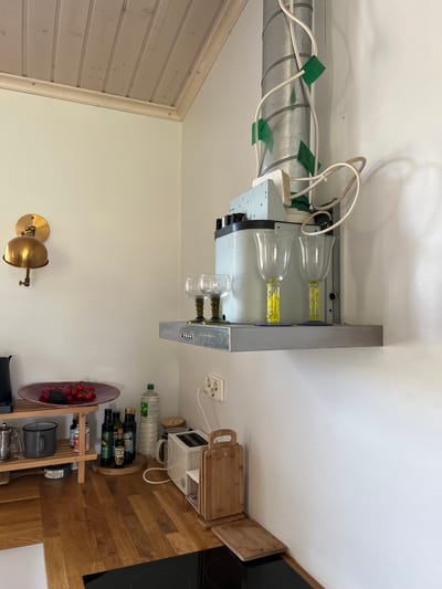 The image depicts a domestic kitchen in Hämeenlinna, Finland. The scene is a close-up of a corner area, focusing on a vent hood, a shelf, and the countertop below. The vent hood is a utilitarian, metallic structure with a large, corrugated pipe ascending the wall. White electrical cords are taped to the pipe with green tape. Below the pipe, a grey metal shelf holds three wine glasses with decorative yellow stems, two of which are more prominent in the foreground. The shelf itself is mounted on the wall beneath the vent hood unit. To the left of the vent hood, a brass wall lamp with a bell-shaped shade is visible. Below the shelf and lamp, a wooden countertop is laden with various items. A maroon plate holds a cluster of cherry tomatoes and an avocado. To the right of the plate is a tiered wooden shelf unit holding kitchenware, including a grey mug and a glass carafe. Further to the right on the countertop are several bottles of olive oil and other condiments, a white toaster, and a wooden cutting board. Electrical outlets are present on the wall to the right of the shelf. The overall environment suggests a functional, lived-in kitchen space.