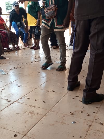 The image displays an active, covered outdoor or semi-enclosed public area, located in the city of Ajalli, Nigeria, characterized by a light-colored tiled floor heavily littered with dark brown, organic-appearing debris, resembling groundnut shells, interspersed with white plastic waste.
Five individuals are visible. On the far left, a person is seated, wearing a red top and a patterned maroon garment on their lower body, with light-colored sandals on their feet. Adjacent, a woman wearing a black cap, black short-sleeved top, and blue jeans leans forward, carrying a dark shoulder bag and wearing light-colored open-toed shoes. Behind her, a person in a green and yellow jersey holds a black handbag and wears red flat shoes. In the central foreground, a person in a green and white patterned tunic, grey jeans, and green sandals is captured mid-step, their right foot elevated. This individual carries a brown shoulder bag with a distinct monogram pattern worn across their chest. On the far right, the lower legs of a person wearing dark brown trousers and black closed-toe shoes are visible, indicating a standing posture. The scene is brightly lit, casting subtle shadows on the tiled surface.