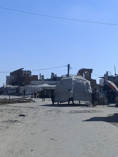 The image depicts a street scene in what appears to be a damaged urban area, possibly Gaza. In the foreground, a dirt or gravel road is visible, with scattered debris. A few people are walking on the road, casting shadows.
In the middle ground, several structures are present. Some appear to be makeshift tents or covered stalls, possibly for market activity. One large structure is covered with a gray tarp, creating an irregular, tent-like shape. Behind these, more substantial buildings are visible, some of which are clearly damaged or in ruins, with broken walls and exposed interiors. One building has a large arched opening at the top.
Several individuals are scattered throughout the scene, some walking, others standing near the stalls or buildings. They are dressed in casual clothing. The presence of tents, stalls, and people suggests some level of activity, possibly a market or a temporary settlement, despite the surrounding destruction. An umbrella, possibly an orange and gray one, is visible to the right. A white van is parked behind some of the tents on the left.
The sky is clear and blue, indicating a sunny day, likely mid-morning or afternoon. The overall impression is one of resilience and ongoing life amidst devastation. No specific text is clearly legible in the image.