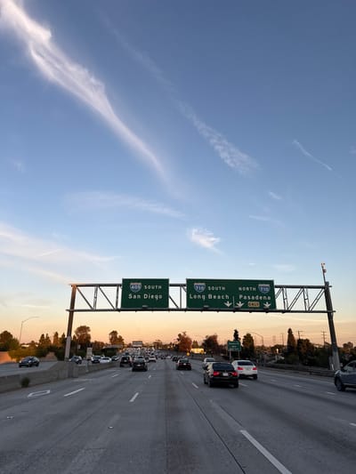 A highway scene under a blue sky with wispy clouds. Traffic is visible in the right lanes. Overhead road signs direct drivers to destinations: 405 South towards San Diego, 710 South towards Long Beach, and 710 North towards Pasadena. Trees line the edge of the highway. The horizon shows a warm sunset glow.