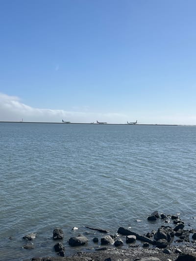 An eye-level, medium shot captures a calm, wide expanse of grey-blue water in the foreground, with a dark, rocky shoreline visible at the very bottom of the frame. In the middle ground, a thin, flat strip of land stretches horizontally across the image, appearing to be a runway or causeway. Three airplanes are visible on this strip of land. On the far left, an airplane is positioned on the land, with a tall, slender structure resembling a control tower or navigational beacon behind it. To the right of that, a second airplane is also on the land. Further to the right, a third airplane is visible. The background consists of a clear, pale blue sky with wispy white clouds forming a band across the horizon behind the landmass. The scene suggests an airport located near the water, possibly San Francisco International Airport (SFO) given the proximity to Burlingame, California, which is bordered by the San Francisco Bay.