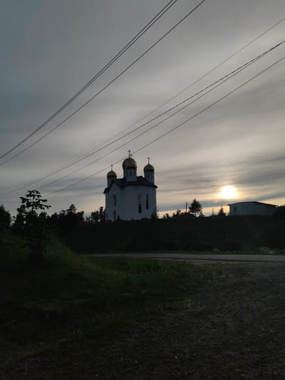 The image depicts an exterior view of a white church with golden domes, situated on a hill overlooking a wooded area. The church has a tall, central structure with multiple smaller domes and a cross on each. The sky is overcast with streaks of light, suggesting either sunrise or sunset, with the sun partially visible as a bright orb. Several power lines are strung across the upper portion of the frame, running diagonally from left to right. In the foreground, there is a gravel path or road and a patch of dark grass. To the right of the church, a low-rise building with a light-colored roof is visible. The scene is devoid of any people or discernible activity. The overall impression is one of a quiet, rural landscape with a prominent religious structure.