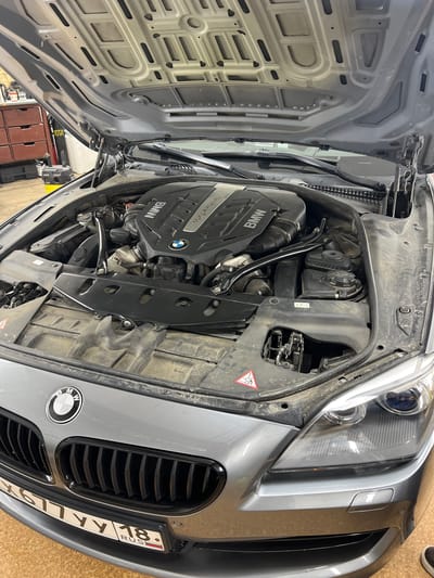 A gray BMW 6 Series (F13/F12) coupe is parked indoors, likely in a garage or workshop. The hood is open, revealing the V8 twin-turbo engine. The car's front end, including the grille and headlights, is visible. A Russian license plate, "X677YY 18," is mounted on the front bumper. The environment appears to be a workshop, with tools and equipment visible in the background, such as metal cabinets and a vacuum cleaner. The lighting suggests an indoor setting. There are no people visible in the image.