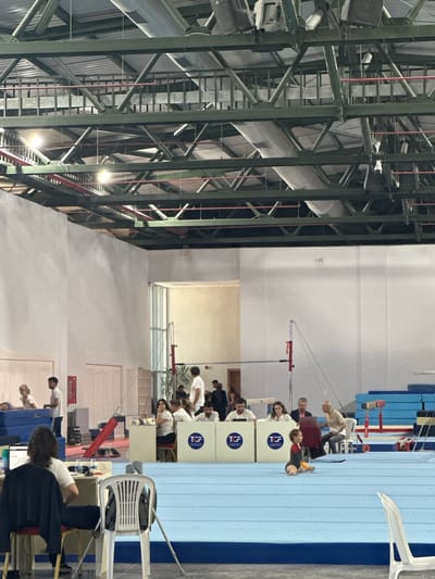 This image depicts a gymnastics competition or training session within a large indoor facility in Sancaktepe, Turkey. The scene is characterized by a high ceiling with exposed industrial lighting and ductwork. In the foreground, a blue gymnastics mat covers a significant portion of the floor. A young girl in a leotard is sitting on the mat, exhibiting a straddle split.

Several people are visible in the middle ground, positioned behind a series of white partitions with "TCF" logos. These individuals appear to be officials or judges, some interacting with laptops or other equipment. A few athletes, possibly waiting for their turn or observing, are also present in this area. Further back, individuals are seen near stacked blue mats, suggesting equipment storage or preparation.

The background shows more of the expansive gymnasium, with stark white walls and the continuation of the industrial ceiling structure. The lighting in the facility appears to be a mix of overhead fluorescent and spotlights. The overall environment suggests a professional or semi-professional sporting venue.