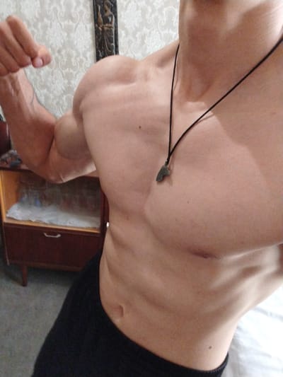 The image shows a close-up of a muscular person's torso, flexing their bicep. They are wearing a black necklace with a pendant and dark clothing. In the background, there is a dresser and patterned wallpaper. The image was taken indoors, in what appears to be a domestic setting. There is no indication of the city of Kliment, Bulgaria, or any other specific location details.