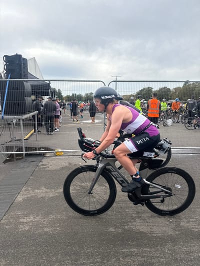 The image displays an outdoor triathlon event in Weymouth, United Kingdom, under an overcast sky. In the foreground, a female cyclist, identifiable by race number 2290, is depicted in profile, riding a grey Felt aerodynamic triathlon bicycle equipped with black Hunt deep-section wheels. She wears a black KASM helmet, a purple and white sleeveless triathlon suit with "DUTI" on the shorts, and black cycling shoes. A watch is visible on her left wrist. She maintains an aerodynamic riding posture.
Metal crowd barriers bisect the scene horizontally. To the left of the cyclist, a multi-tiered stack of black professional audio speakers is mounted on a grey metal scaffolding structure, positioned adjacent to a partially visible white tent. Several individuals, including one in a high-visibility yellow jacket, stand behind these barriers. The ground surface in the foreground is wet, dark asphalt, with a yellow object visible near the speaker setup.
To the right of the cyclist and behind the barriers, a crowded transition area is visible, populated by numerous individuals in athletic attire or high-visibility orange and yellow vests, alongside many parked bicycles. Trees line the distant background under the uniform grey sky, and a street light pole stands centrally in the mid-distance. The scene depicts an active, organized sporting event.