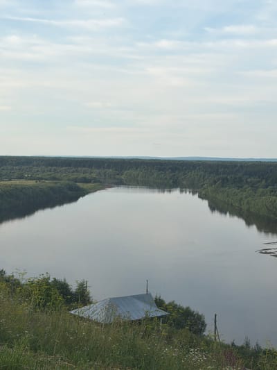 A high-angle, full-shot photograph captures a wide river flowing through a lush, green landscape. The river dominates the center of the frame, its surface reflecting the pale blue sky and scattered white clouds. The riverbanks are densely forested with a variety of trees, creating a verdant wall that stretches into the distance. On the foreground, a small, rustic building with a metal roof sits nestled amidst tall grasses and wildflowers, suggesting a rural or remote setting. The scene appears serene and untouched, evoking a sense of natural beauty and tranquility. The location is identified as Cherdyn, Russia.