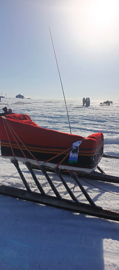 The image displays a vast, snow-covered landscape under a bright, clear sky, consistent with a winter environment in Nelmin-Nos, Russia.

In the foreground, a traditional wooden sled is positioned, angled from the lower left towards the mid-right. The sled's runners are dark, likely wood, and appear to be frosted. The main body of the sled holds cargo, prominently covered by a thick, red, quilted fabric. Below the red covering, a section of patterned fabric is visible, featuring horizontal stripes in black, orange, and light green. Several ropes, colored green, orange, and light blue, crisscross the cargo, securing it to the sled frame. A small, rectangular white fabric banner with a green and blue design is affixed to the right side of the cargo. A thin, dark pole or antenna extends vertically from the sled towards the upper sky.

The mid-ground and background consist of an expansive, flat expanse of snow and ice, exhibiting textured surfaces. To the far left, a dark, low-slung building or shelter is visible on the distant horizon, accompanied by several vertical poles or structures. To the mid-right, a cluster of approximately three to four individuals, dressed in heavy winter clothing, stands together; one figure's back displays the number '21'. Further to the right, a second traditional sled is present, with at least two dark-colored reindeer harnessed to it. The sun, positioned in the upper right quadrant of the sky, casts strong light and shadows across the scene.