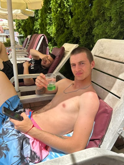 The image depicts two young men relaxing on lounge chairs outdoors, possibly at a resort or pool area, with lush green foliage in the background. In the foreground, a Caucasian man with short hair is seated, holding a clear plastic cup with a green beverage and a mobile phone in his left hand. He is wearing blue swim trunks with a palm tree pattern. To his left, another man, possibly of Middle Eastern descent, is lying down, with his right hand covering his eyes. He is wearing a black t-shirt. The lounge chairs are made of a light-colored material, and there are umbrellas visible further back. The lighting suggests a sunny day. While the provided context mentions Zverino, Bulgaria, the image itself does not contain any explicit visual cues to confirm this location.
