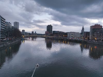 The image displays a wide, overcast urban river scene in Liège, Belgium, likely viewed from a bridge or elevated structure. The time of day appears to be twilight or early evening, characterized by dark, heavy clouds and artificial light sources beginning to dominate.
The Meuse River occupies the central and foreground portions of the frame, exhibiting a dark, reflective surface that mirrors the sky and city lights. In the lower center foreground, a white, triangular object, possibly a navigation marker or part of a structure, extends diagonally out of the water.
Along the left bank (viewer's left), a concrete or stone quay runs parallel to the river. Above the quay, a row of multi-story buildings, including modern residential blocks with numerous windows, stands adjacent to a tree-lined street. The trees are bare, suggesting an autumn or winter season. Several vehicles are visible parked along this street, and at least two boats are moored to the quay.
The right bank (viewer's right) features a mixed architectural landscape, combining older, traditional structures with some contemporary buildings. A prominent, dark-spired building, possibly a church, rises above the mid-ground skyline. A busy road runs along this bank, displaying visible headlights and taillights from numerous vehicles, which create streaks of light reflected on the river. Multiple streetlights illuminate the riverfront.
In the mid-ground, several bridges span the river, connecting the two banks. Some of these bridges are illuminated, their lights casting reflections on the water. The cityscape extends into the distant background on both sides of the river, marked by various building silhouettes against the uniformly grey and cloudy sky. No discernible individuals are visible within the frame.