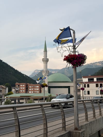 This image depicts a street scene in Konjic, Bosnia and Herzegovina. The primary subjects are flags, a lamppost with a hanging flower basket, a minaret of a mosque, and buildings. The scene is set against a backdrop of a mountainous landscape and a cloudy sky, suggesting it is daytime with potentially overcast weather.
Several flags are visible. Two prominent flags appear to be the national flag of Bosnia and Herzegovina, recognizable by its blue and yellow design with white stars. Another flag, white with blue and yellow accents, is also present, and its specific significance is not immediately clear from the image alone. The flags are attached to a lamppost adorned with a basket overflowing with vibrant pink and red flowers.
To the left, the slender, white minaret of a mosque with a green spire rises, a clear indicator of the religious and cultural context of the location. Behind the minaret, the green dome of the mosque is partially visible. Several multi-story buildings with brick facades and numerous windows are situated on a hillside, interspersed with trees.
A modern, white car is parked on a road that runs alongside a metal railing. The railing, made of curved metal bars, suggests a walkway or elevated position overlooking the road and buildings. The overall impression is one of a town nestled within a valley, with a blend of modern infrastructure and historical architectural elements. No explicit activity or event is taking place; the image captures a static moment. The lighting suggests it is likely late afternoon or early morning. There is no visible text in the image.