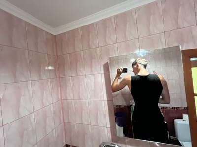 The image depicts a bathroom with pink marble-patterned tiles covering the walls and a white ceiling with decorative molding. In the center of the image, a mirror reflects a person wearing a black sleeveless dress. The person, whose back is to the camera, is holding a phone and taking a selfie. Their hair is short and styled. Visible in the reflection are a blue bucket, a white toilet, and a wooden door frame. The scene appears to be located in the city of Sobishchytsi, Ukraine.