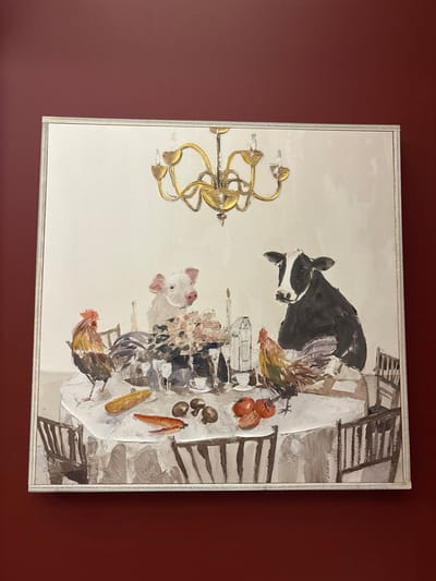 An oil painting of a dining table scene. A black and white cow, two chickens, and a pig overlook the meal. A chandelier hangs to the side. The table is set with tomatoes, mushrooms, carrots, corn, flowers, and bottles. Chairs are positioned around the table. The painting has a textured, three-dimensional effect. The artwork is framed. The wall it hangs on is a deep red color.