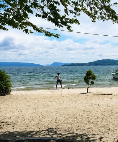 A person stands on a sandy beach facing a body of water. The person is wearing a light-colored shirt and dark shorts, with their arms slightly outstretched. The beach is composed of light-colored sand and some scattered debris. To the right of the person, a small, conical evergreen tree grows. The water is a deep blue-green with visible waves, and in the distance, hills or mountains form the horizon under a cloudy blue sky. A boat with a white hull and green trim is moored on the right side of the water. Overhead, dark green leaves from a tree frame the top of the image. The overall scene suggests a natural, possibly recreational, setting at the edge of a lake or sea. The location is Lumban Bul Bul, Indonesia.