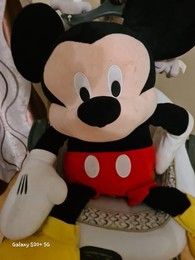 A plush Mickey Mouse toy is the central subject of the image. The toy is large and appears to be sitting on a chair or couch. It is wearing its signature red shorts with white buttons and yellow shoes. The Mickey Mouse toy is dark black with large round ears, a tan face, and white eyes with black pupils. The background of the image consists of what appears to be a patterned curtain on the left and a light-colored surface that the toy is resting on. The image is taken indoors. The lower left corner of the image displays "Galaxy S20+ 5G". There is no indication of the specific location within Woodburn, United States of America.