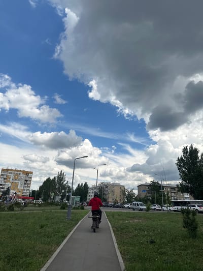 The image depicts a street scene in Lopaskyne, Ukraine, on a cloudy day. A person wearing a red jacket and a helmet is riding a small motorbike along a paved path. To the left of the path is a grassy area, and beyond that, a playground and a multi-story apartment building. On the right side of the path, there is a road with several cars and a bus, and more apartment buildings in the background. Tall trees are visible throughout the scene. The sky is filled with dramatic, grey clouds interspersed with patches of blue sky and wispy white clouds. The overall impression is a typical urban environment on an overcast day.