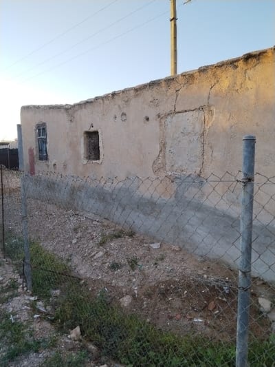 A dilapidated wall with two boarded-up window openings is lined by a chain-link fence. Patches of green vegetation are visible below the fence, and a utility pole rises in the background. The wall is beige, with visible cracks and deterioration. The ground is dirt and gravel, showing signs of neglect. The scene suggests a state of abandonment or disrepair.