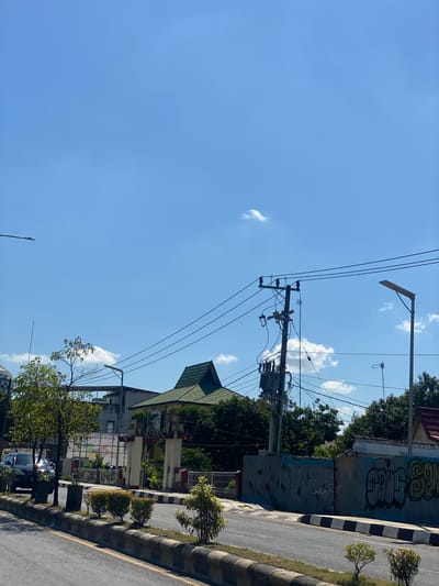 A street-level, eye-level shot shows a clear blue sky with a few small clouds over a road in a city in Jawa, Indonesia. A power pole with a transformer and multiple wires dominates the center-right of the frame, with a street light visible to its right. The road is bordered by a median strip with small trees and shrubs. Beyond the median, a fence covered in graffiti runs along a property line, with a building featuring a distinctive green, peaked roof visible behind it. A car is parked on the left side of the road, and a tall, thin pole is positioned near a leafy tree on the left side of the image. The overall impression is of a typical urban street scene on a sunny day.