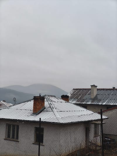 The photograph captures a section of Borino, Bulgaria, on a cloudy, overcast day. The scene is dominated by rooftops covered with a light dusting of snow. The closest building is a single-story structure with a white exterior and a metal roof, featuring two brick chimneys. The building is partially obscured by a chain-link fence. To the rear, another building with a weathered metal roof and a white chimney is visible. Beyond the buildings, hazy mountains are silhouetted against the gray sky. The foreground includes dry vegetation poking through the bottom of the fence. No people are visible.