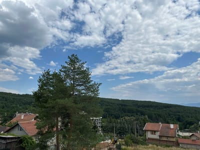 The image depicts a landscape scene in Pchelin, Bulgaria. In the foreground, there are tall pine trees. Behind the trees, a cluster of houses with red tiled roofs is visible. Further in the background, a dense, dark green forest covers a hillside, and a layered mountain range can be seen in the distance. The sky is a bright blue with scattered white and gray clouds. A small yellow excavator is parked on a construction site in the middle ground. There are no people or animals present in the image.