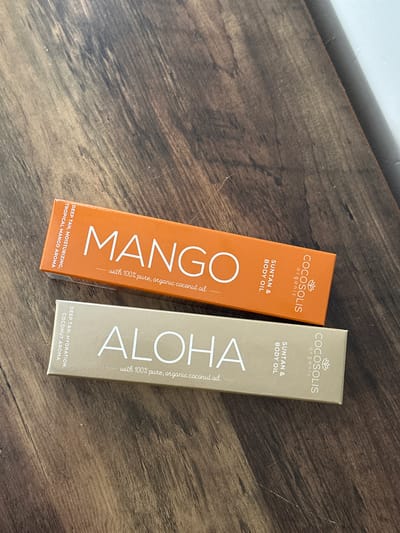 Two rectangular boxes of Cocolis organic suntan and body oil are displayed on a wooden surface. The top box is orange with the word "MANGO" printed in large white letters, along with the text "with 100% pure, organic coconut oil" in smaller white letters. The left side of the orange box has vertical text that reads "DEEP TAN, MOISTURIZING, TROPICAL MANGO AROMA". The right side of the orange box has vertical text that reads "COCOSOLIS ORGANIC" and "SUNTAN & BODY OIL".

The bottom box is gold with the word "ALOHA" printed in large white letters, along with the text "with 100% pure, organic coconut oil" in smaller white letters. The left side of the gold box has vertical text that reads "DEEP TAN, HYDRATION, COCONUT AROMA". The right side of the gold box has vertical text that reads "COCOSOLIS ORGANIC" and "SUNTAN & BODY OIL".

The image does not contain any information to determine the location in Belfast City, United Kingdom.