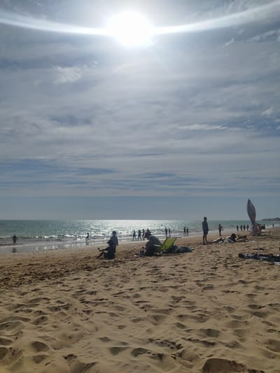 This media file depicts a sunny day at the beach in Rota, Spain. The scene is set on a sandy shore with gentle waves rolling in from a sparkling turquoise ocean. The sun is high in the sky, creating a bright, almost hazy atmosphere with a lens flare visible in the upper portion of the image. A few scattered clouds drift across the sky, indicating fair weather.

Several people are enjoying the beach. Some are wading or playing in the shallow water, while others are relaxing on the sand. In the foreground, a person is sitting on a beach chair. Further back, a group of people are walking along the shoreline, and others are scattered across the beach, some lying down, suggesting a leisurely atmosphere. A colorful beach umbrella stands in the middle ground, adding a touch of vibrancy to the scene. The overall mood is one of relaxation and enjoyment of a summer day. The time of day appears to be midday or early afternoon, given the brightness of the sun and the angle of the light.