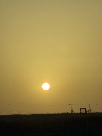 The image captures a serene landscape during either sunrise or sunset, presenting a warm, golden-yellow sky that occupies the majority of the frame. The sky is largely uniform, with a brighter, more intense yellow radiating from the sun. The sun itself is a prominent, bright white-yellow disk, positioned off-center towards the right.

In the lower third of the image, a dark, silhouetted band forms the horizon, suggesting a field or low-lying vegetation. Stretching horizontally across the frame, just above this dark landmass, are multiple power lines. These lines feature several evenly spaced, small, dark spherical objects, likely bird diverters or line markers.

Two distinct industrial structures, appearing as transmission towers, are visible in silhouette against the bright sky. The tower on the left exhibits a classic lattice design. Between these two towers, a dark, rectangular structure, possibly part of an electrical substation or a fence, is also silhouetted. No people are visible, and there is no discernible text within the image. The scene evokes a sense of vastness and tranquility, with the golden hour light illuminating the utility infrastructure against the natural backdrop.