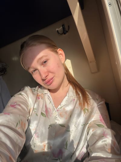A woman is lying down in what appears to be a train compartment, wearing floral pajamas. The location of the scene is unknown, and it is not possible to determine if the image was taken in Krasnyy Sulin, Russia. The train compartment has a shelf, a hook, and a window allowing sunlight to enter.