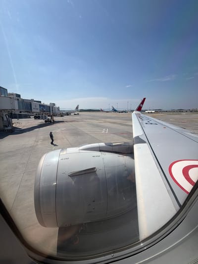 The image provides a view from an airplane window, looking out onto an airport tarmac on a bright, clear day. The large, white wing of the aircraft, bearing the registration "B-322K," extends across the upper right portion of the frame. Below the wing is a prominent engine nacelle, with "NO STEP" warnings clearly visible in several places.

On the concrete tarmac, a jet bridge is connected to the aircraft. Text on the jet bridge includes numbers such as "16," "911," "90X," and "90Y," alongside some Chinese characters. A single ground crew member, dressed in dark clothing, is walking on the tarmac near the engine, casting a long shadow, suggesting either mid-morning or late afternoon. In the distance, other airplanes are parked at gates, including one with a distinctive red tailfin.

The sky is a clear, bright blue, indicating sunny weather. The scene depicts a typical busy airport environment, with the aircraft parked at a gate, possibly preparing for departure or having recently arrived. Given the visible aircraft registration and Chinese text, the location is consistent with an airport in Jianyang City, China.