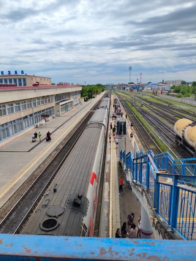 This is an overhead shot of a train station in Buzuluk, Russia. The scene depicts a moderately busy train station with several tracks, a train stopped at the platform, and a station building. People are visible on the platform, some with luggage, suggesting they are travelers. The weather appears cloudy, and the time of day seems to be late morning or early afternoon, given the lighting. The station building has a long facade with many windows and a red roof. Blue lettering on the building's facade reads "КЗАА" and "КАЗА". A pedestrian overpass with blue railings is visible in the foreground, with people gathered near its base. In the background, there are more tracks, industrial-looking buildings, and trees. A yellow cylindrical tank car is parked on a track in the distance.
