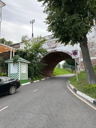 In Voksherino, Russia, on a cloudy day, a black sedan is parked on the left side of a paved road that curves under a brick archway. To the left of the road, a small, green-and-white striped structure resembling a guardhouse or ticket booth stands next to a building with an outdoor seating area. Lush green foliage covers the area around the booth and climbs up the archway. A traffic sign indicating a tow-away zone is posted on the right side of the road, near a large tree with rough bark. The road has white and yellow lane markings and a black-and-white striped curb. The archway reveals a grassy slope and more trees beyond.