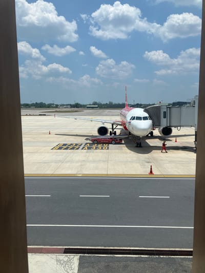 The scene depicts an airplane parked at an airport gate, observed from an elevated position indoors, possibly a waiting area. The airplane is a red and white Airbus A320, with "B737 A320" and "B777 A330" painted on the tarmac near the nose of the plane. A jet bridge is connected to the plane. Two ground crew workers in red uniforms are positioned near the engines. Yellow lines mark the parking area. Further away, the airport grounds extend to the horizon, with trees and buildings. Beside the plane is the asphalt road. The sky is visible in the background, filled with fluffy white clouds against a blue sky. The image appears to be taken at or near Tambon Hua Toei, Thailand, because the plane is a red and white AirAsia Airbus A320, which has hubs in the region.