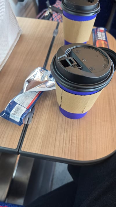 The image shows a close-up of two paper coffee cups with black lids and blue insulating sleeves, resting on a light-colored wooden table. One cup is slightly closer to the foreground and its lid is visible, displaying the text "CAUTION CONTENTS HOT". The other cup is further back. A crumpled wrapper, likely from a snack or confectionery, lies on the table between the cups. The wrapper has some text and graphics, including a barcode and what appears to be nutritional information. In the background, a dark blue fabric is visible, possibly part of a seat or bag. The lighting suggests it's daytime, and the scene appears to be indoors, possibly in a cafe or on public transport. The overall impression is casual and everyday.
