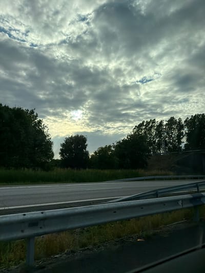 The image captures a cloudy sky over a rural landscape. A highway with guardrails and grassy shoulders is visible in the foreground, suggesting the location is near a road. Trees line the horizon, with the sun partially obscured by clouds, creating a diffused light. There are no people or specific actions depicted, and the scene appears to be a snapshot of a natural environment alongside a transportation route.